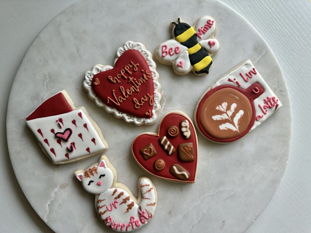 Assorted decorated cookies with Valentine’s Day designs, including hearts, a bee, a coffee cup, and a cat, arranged on a round plate.